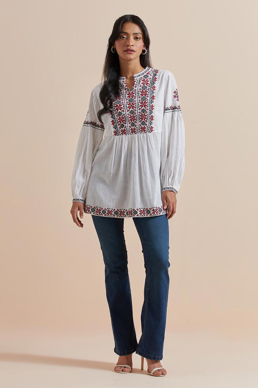 A woman wearing a white cotton embroidered top with a V-neck collar, balloon sleeves, and geometric floral embroidery. She is also wearing earrings and blue jeans.