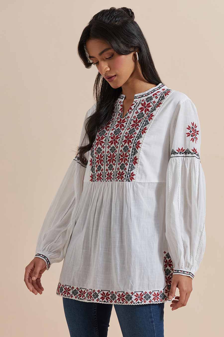 A woman wearing a white cotton embroidered top with a V-neck collar, balloon sleeves, and geometric floral embroidery. She is also wearing earrings and blue jeans.