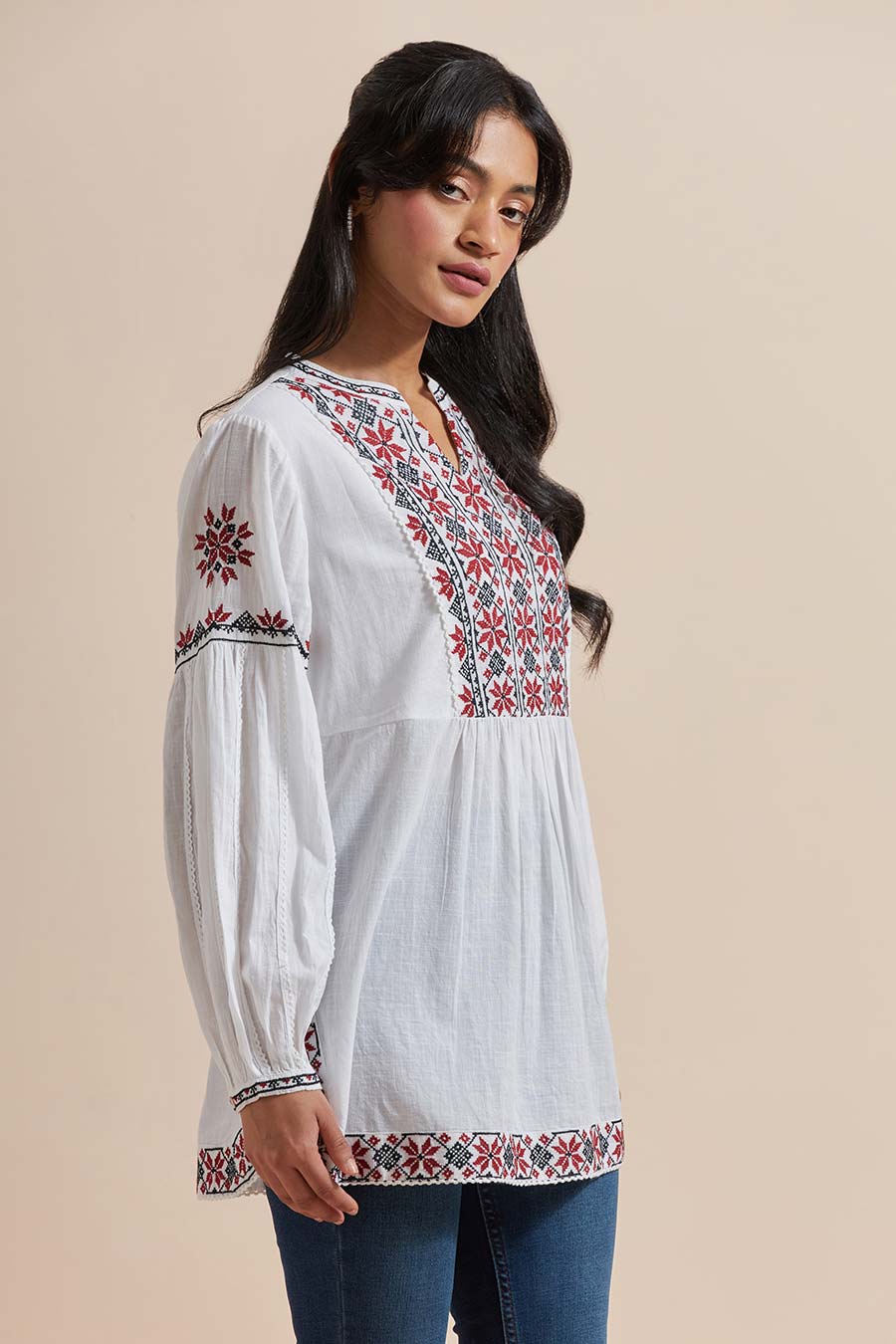 A woman wearing a white cotton embroidered top with a V-neck collar, balloon sleeves, and geometric floral embroidery. She is also wearing earrings and blue jeans.
