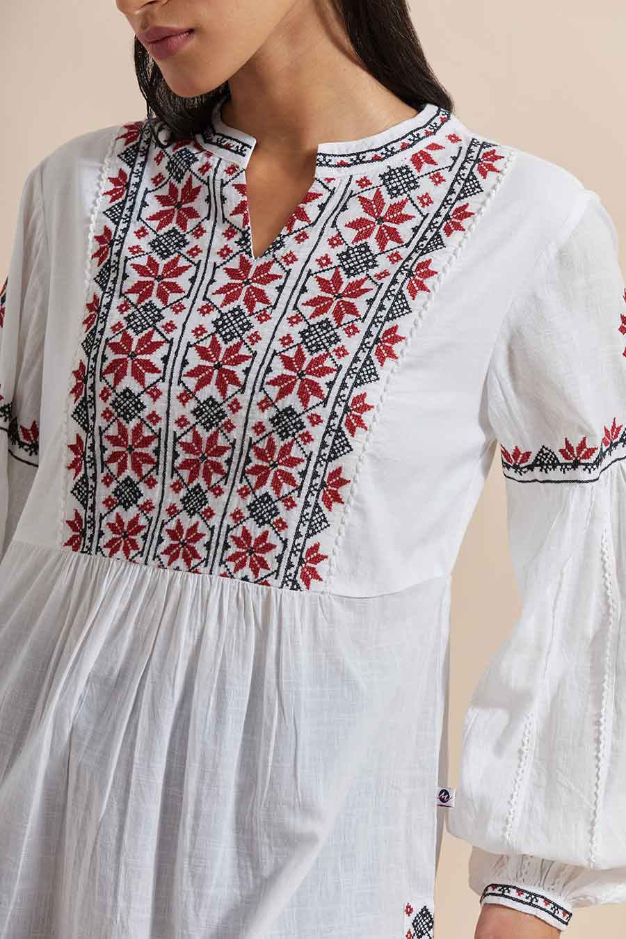 A woman wearing a white cotton embroidered top with a V-neck collar, balloon sleeves, and geometric floral embroidery. She is also wearing earrings and blue jeans.
