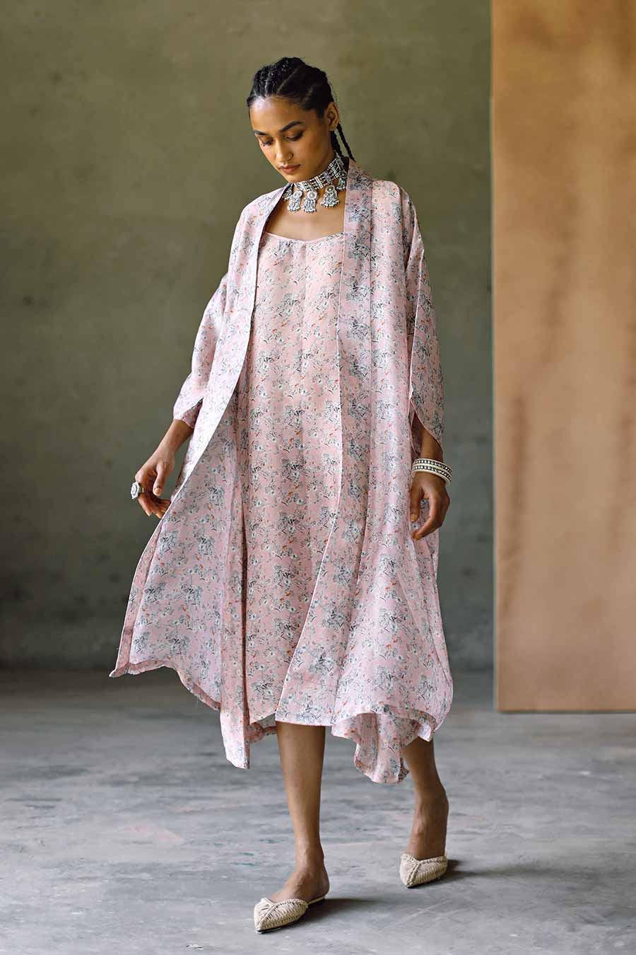 A model wearing a peach-colored floral slip dress with a matching cape, standing in a relaxed pose.